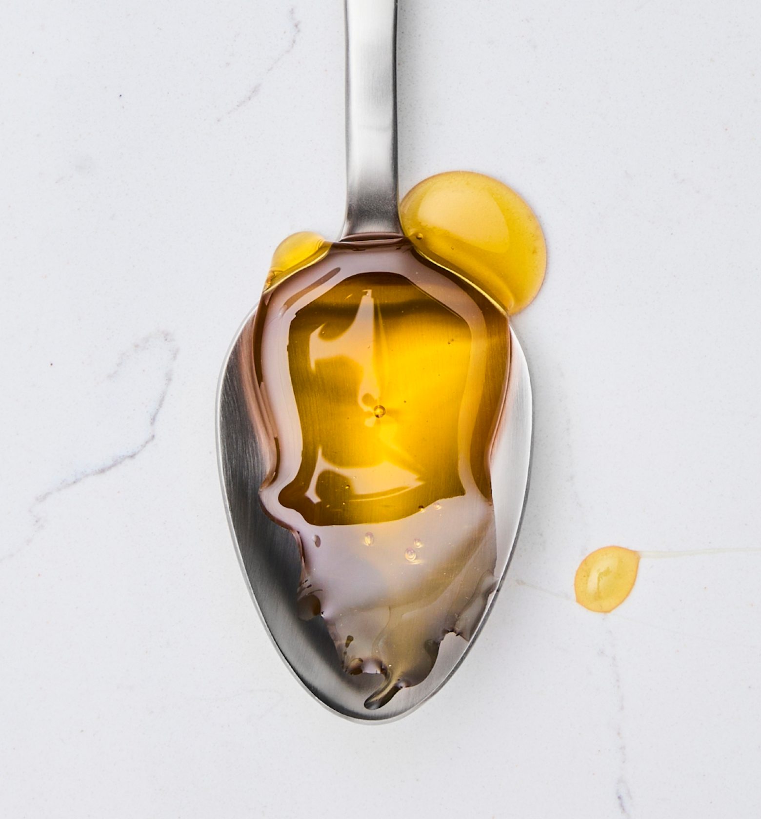 Golden honey or olive oil drizzled on a stainless steel spoon on marble surface